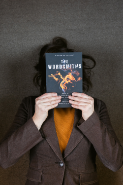 Woman wearing an orange shirt and a brown blazer lying on the floor, holding a speculative fiction anthology book called The Wordsmiths in front of her face