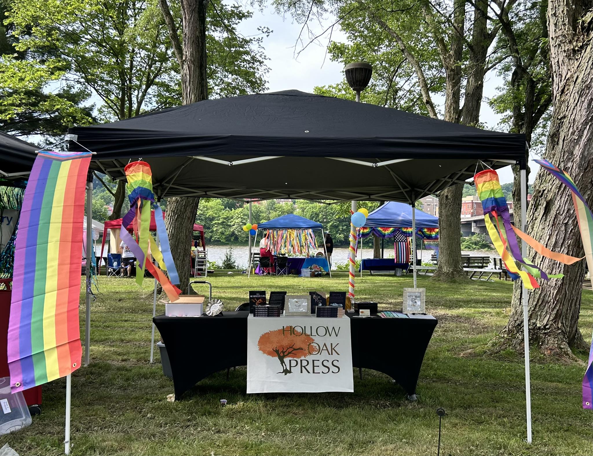 Vendor table under a black tent with rainbow decorations on the sides in a park surrounded by trees